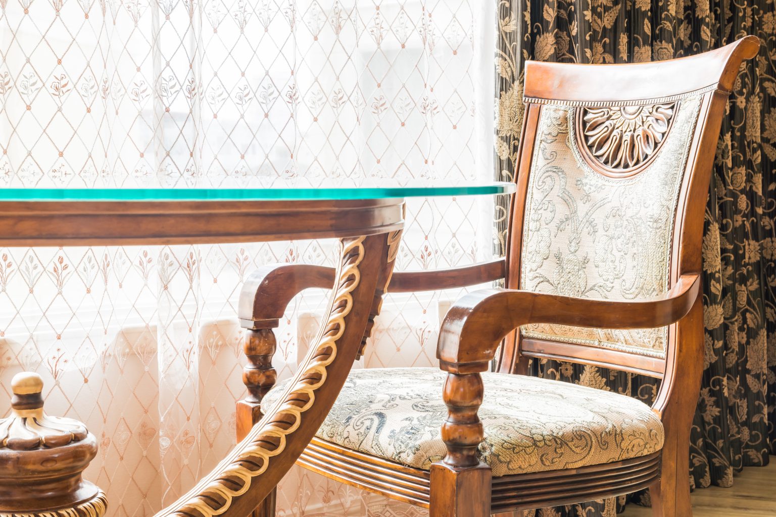Fauteuil en bois sculpté avec un dossier orné et un tissu à motifs floraux, placé à côté d'une table en verre et bois élégante, dans une pièce lumineuse avec des rideaux brodés, idéal pour un tapissier décorateur travaillant sur des projets de meubles classiques et raffinés.