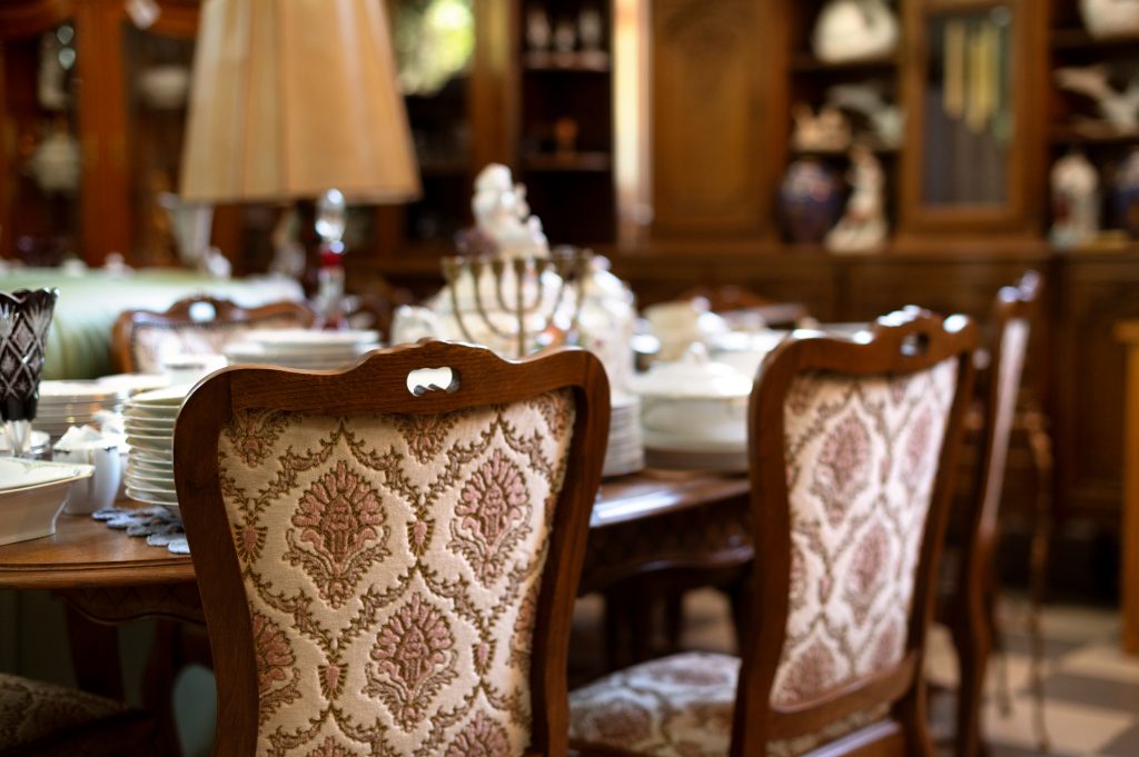Salle à manger élégante avec des chaises en bois ornées de tissu à motifs floraux rose et beige, autour d'une table dressée avec de la vaisselle et des décorations vintage, dans un intérieur riche en bois et objets décoratifs, parfait pour un tapissier décorateur travaillant sur des projets classiques.