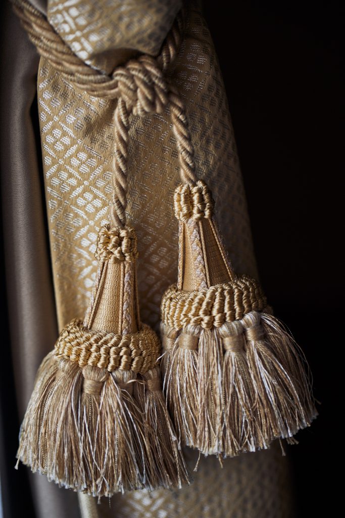 Gros plan sur des embrasses de rideau en corde torsadée beige avec des pompons ornés, attachées à un tissu luxueux à motifs géométriques dorés, parfait pour un tapissier décorateur travaillant sur des projets de décoration intérieure élégants.