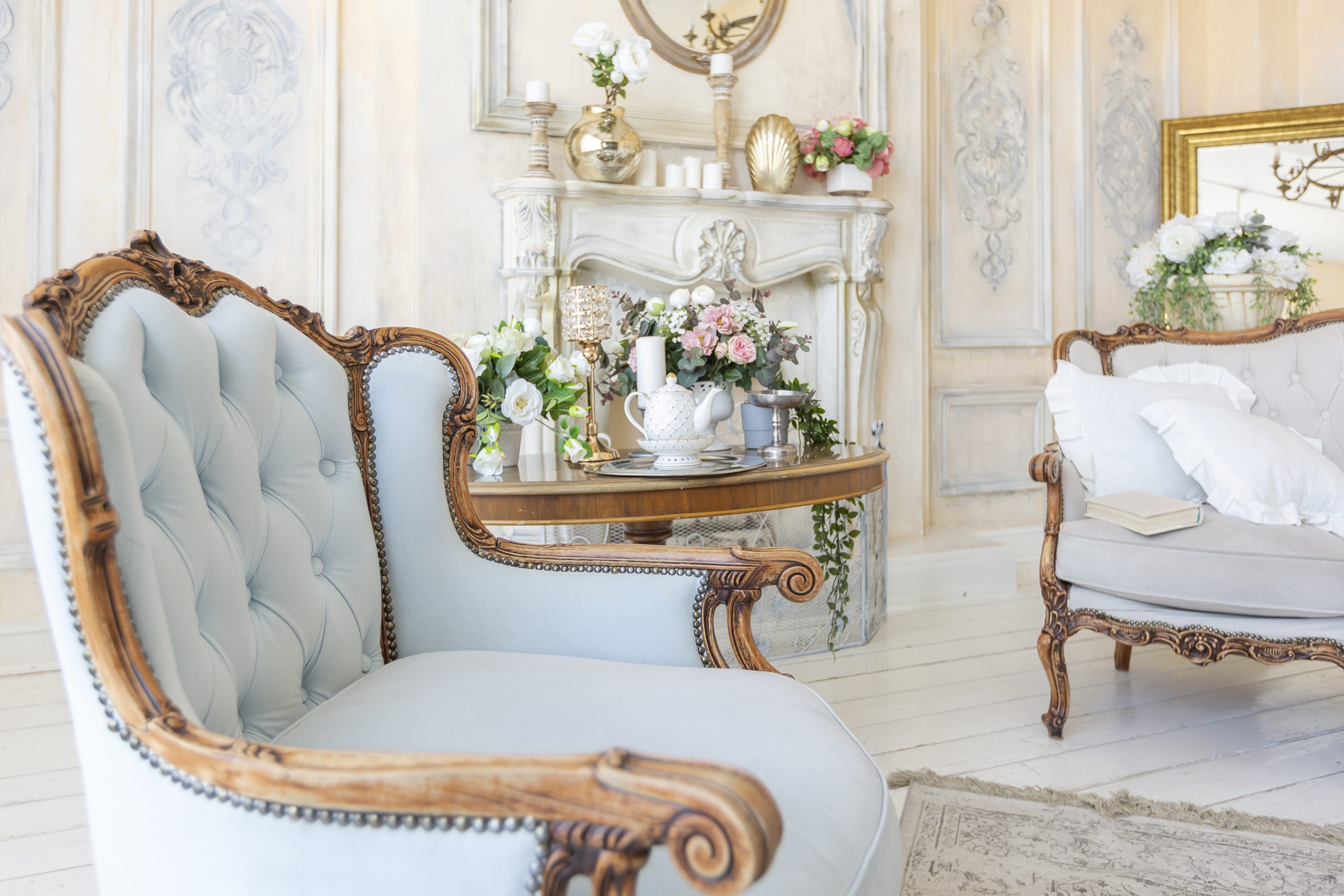 Salon élégant avec un fauteuil capitonné bleu clair à armature en bois sculpté, une table ronde ornée de fleurs et d'objets décoratifs, et un canapé assorti avec des coussins, devant une cheminée ornée, idéal pour un tapissier décorateur travaillant sur des intérieurs classiques et raffinés.