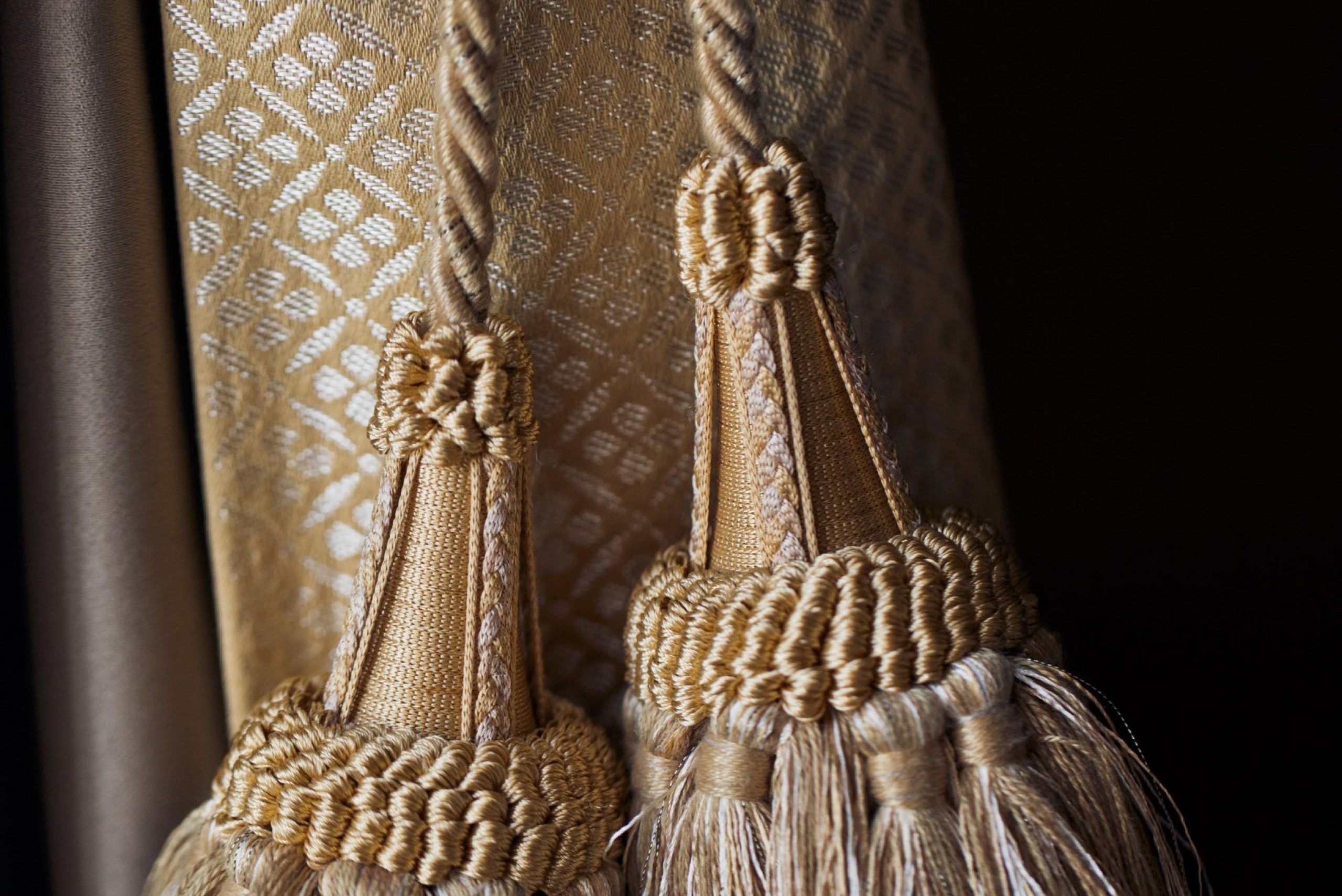 embrasses-rideaux-decor-fenetres Gros plan sur des embrasses de rideau en corde torsadée beige avec des pompons ornés, attachées à un tissu luxueux à motifs géométriques dorés, parfait pour un tapissier décorateur travaillant sur des projets de décoration intérieure élégants.